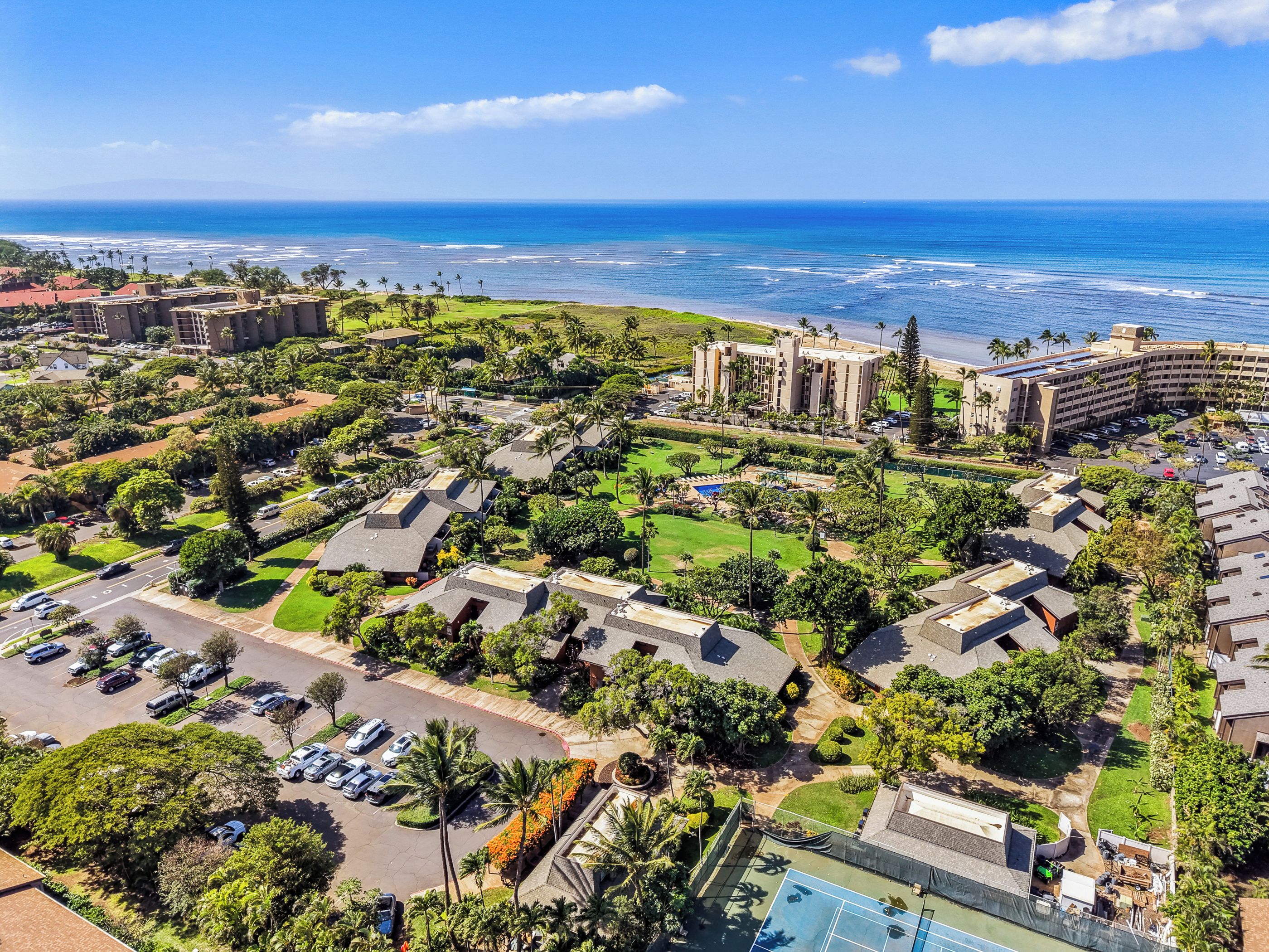 An aerial view of a resort with multiple buildings, a parking area, and a view of the ocean.