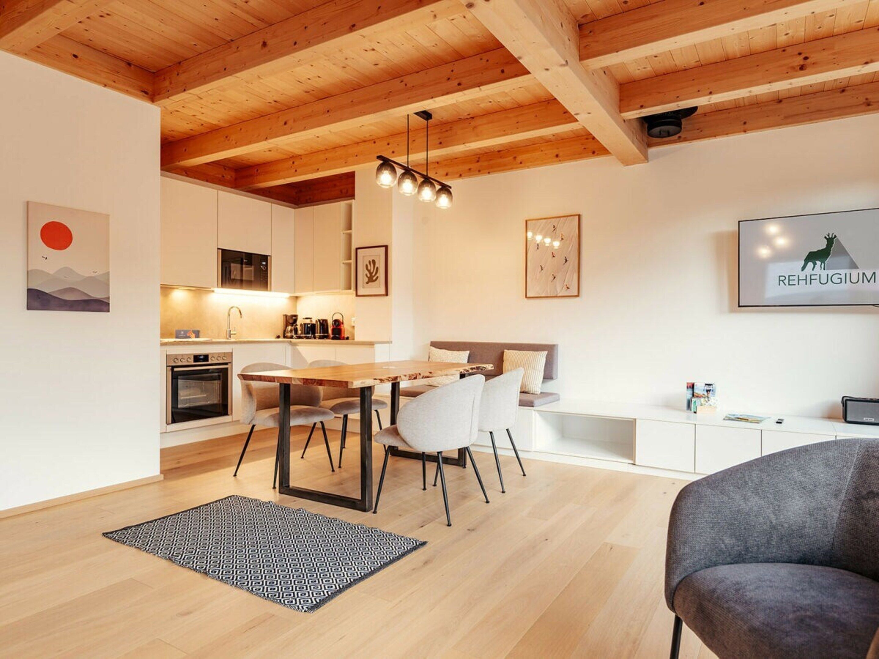 A modern interior with a wooden ceiling, a kitchen with white cabinets, a dining area with a wooden table and chairs, and a living space with a grey sofa.