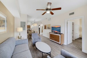 A hotel room with a ceiling fan, a TV on a wooden cabinet, a sofa, a dining table, and a kitchen area in the background. - Magical 2BR Disney Condo - Free Resort Access (Orlando)