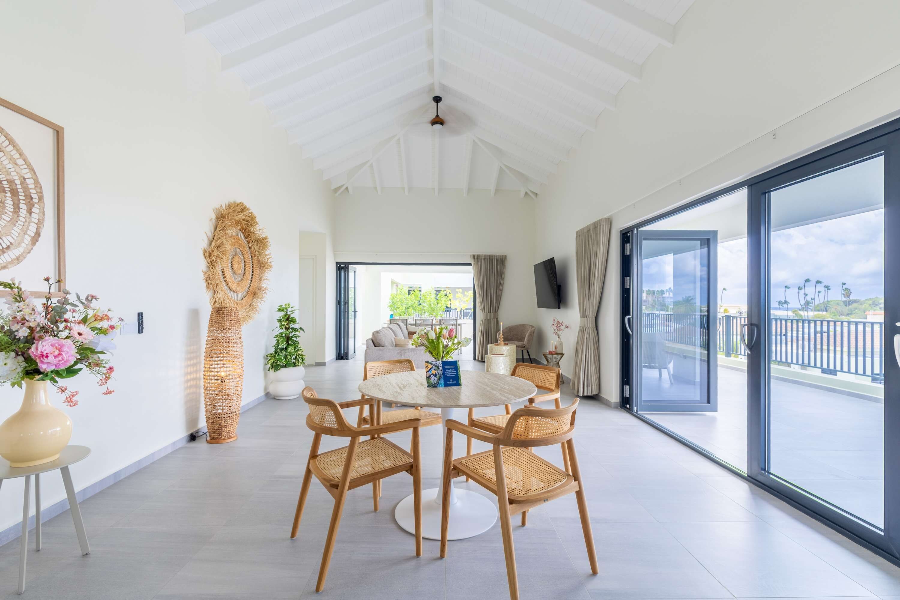A spacious room with a high ceiling, a round dining table with chairs, a large window with a view, and a decorative wall piece.