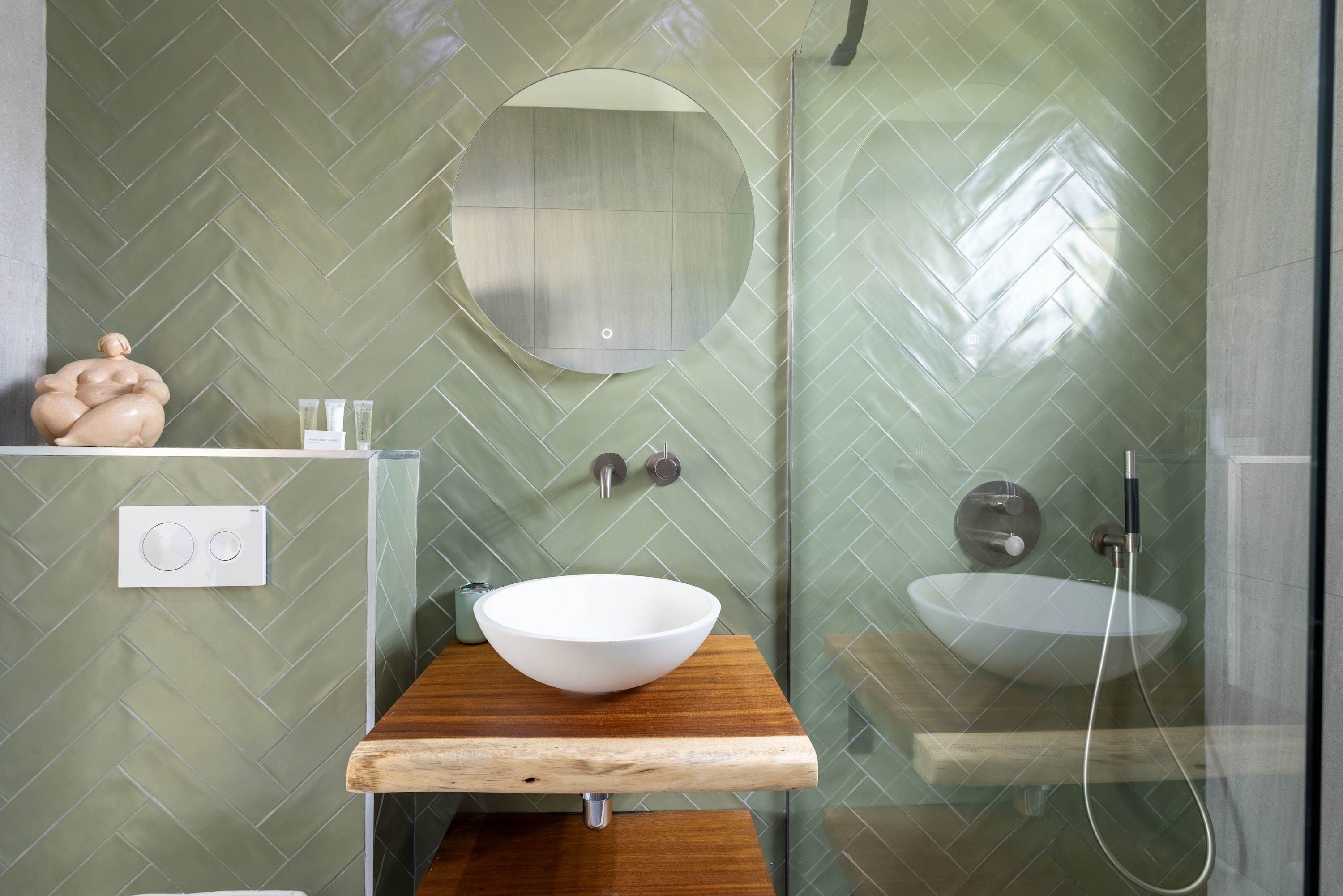 A bathroom with a wooden vanity, a round mirror, a white sink, and a shower.