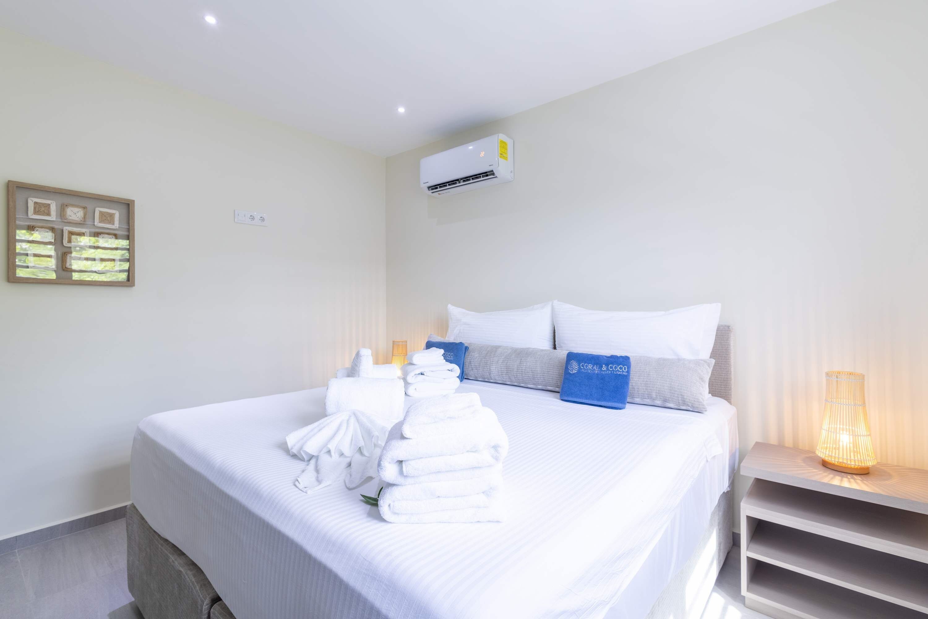A hotel room with a bed, towels, a nightstand, a lamp, and an air conditioning unit.