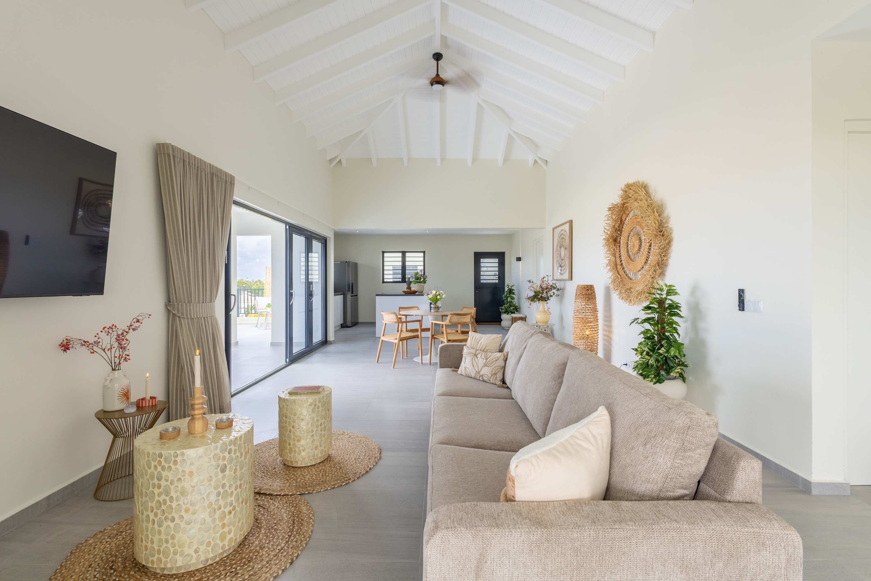 A spacious living area with a sofa, a dining table, and chairs, a kitchen in the background, and a large window with curtains.