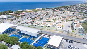 A coastal residential area with multiple-story houses, a main road, and a large sports complex with blue courts. - Altura Luxury 3BR Penthouse by Mambo Beach (Willemstad)
