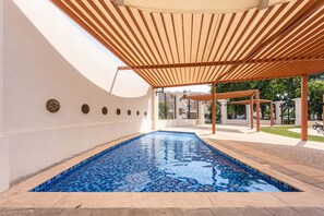 A swimming pool with a tiled floor, surrounded by a covered area with wooden beams and a white wall with decorative elements. - Stayshort - Gorgeous Studio Apartment in Motor City Sleeps 2 With Balcony (Dubai)