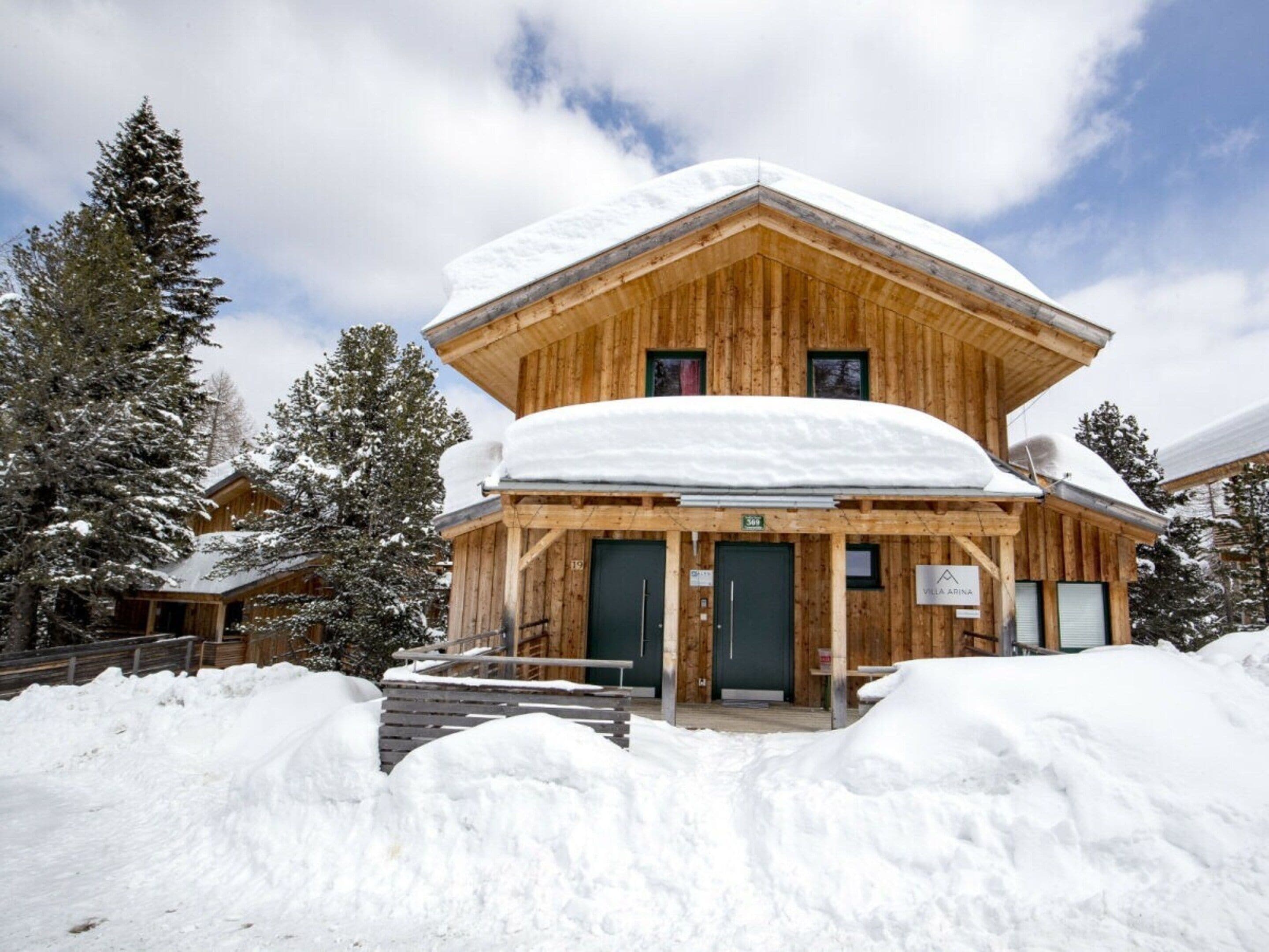 Eine Holzhütte mit schneebedecktem Dach, zwei grünen Türen und einem Schild mit der Aufschrift 'Villa Arina'.