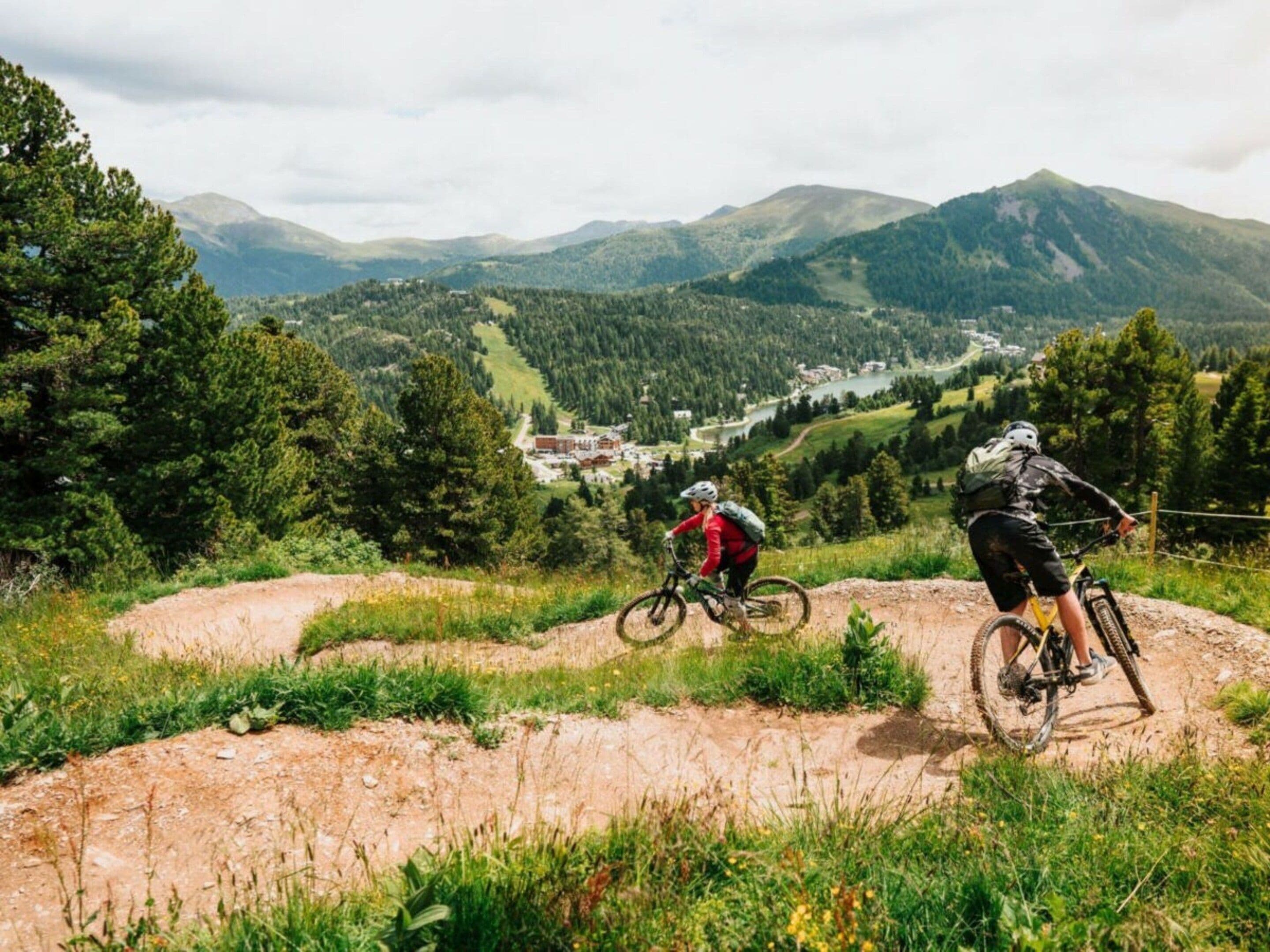 To mountainbikere på en grussti med udsigt over en dal og bjerge i baggrunden.