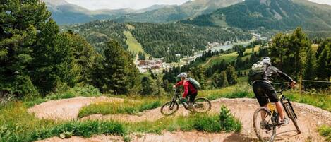 Twee mountainbikers op een onverharde pad met een uitzicht op een vallei en bergen op de achtergrond.
