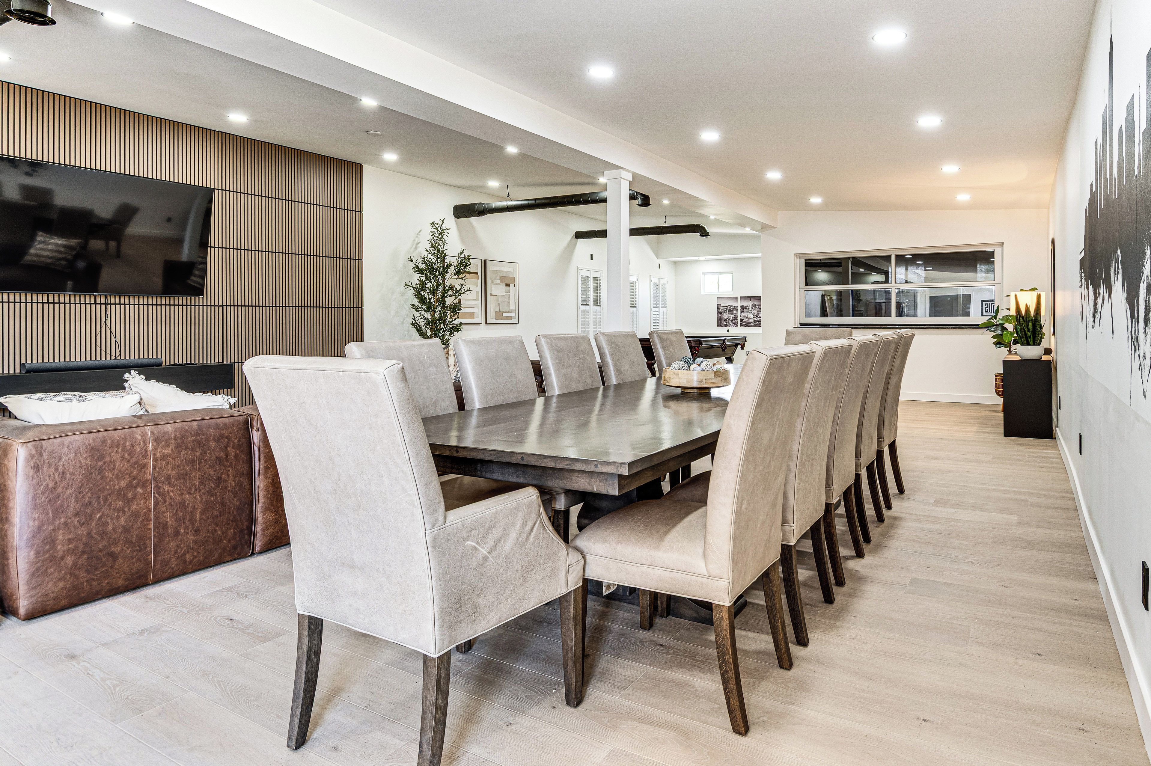 A modern dining area with a long table and multiple chairs, a television mounted on the wall, and a potted plant.