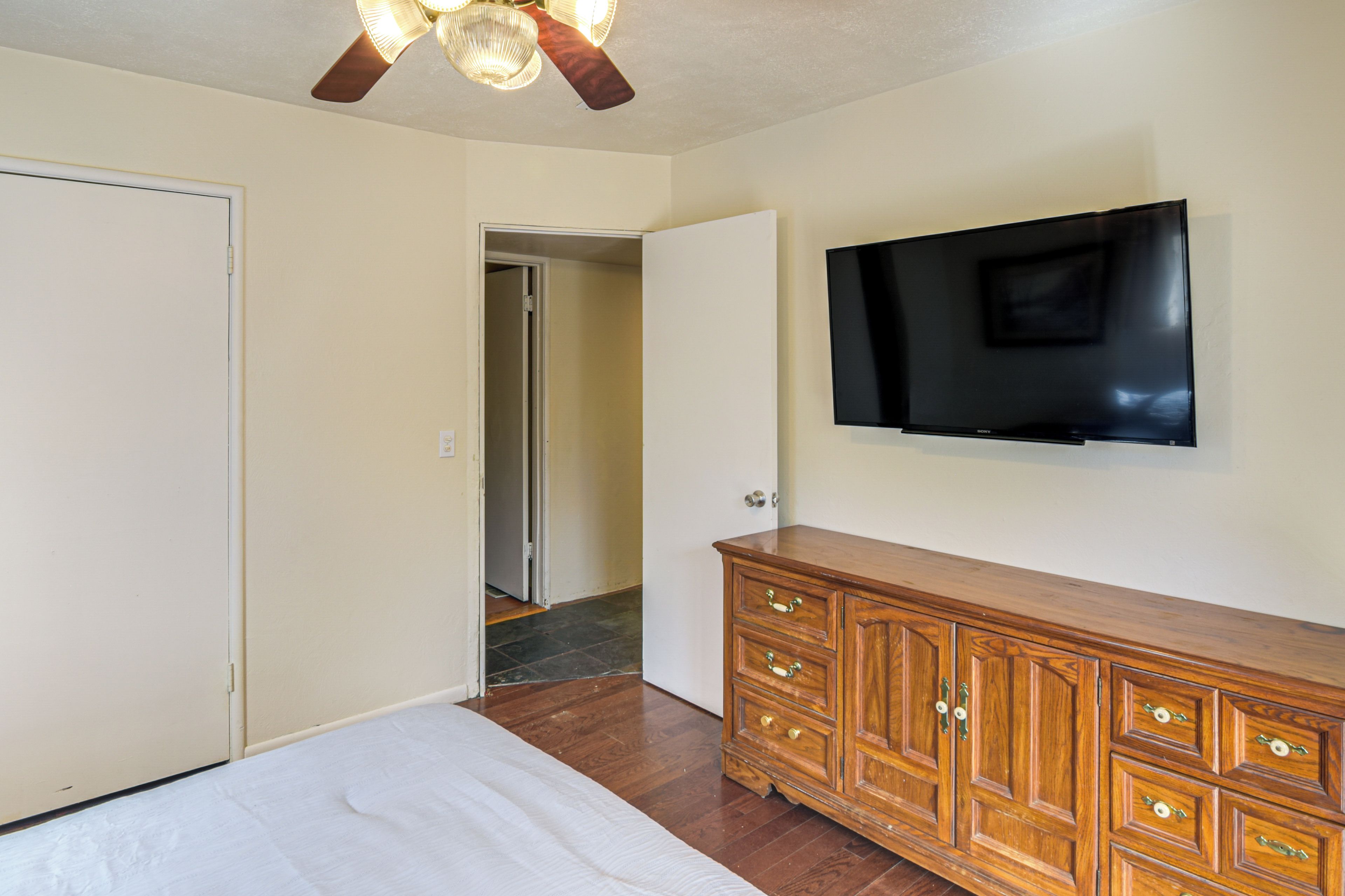 Un dormitorio con cama, cómoda de madera, televisor de pantalla plana en la pared y ventilador de techo.