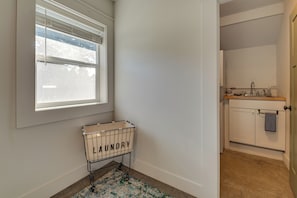 Una cesta de ropa con la etiqueta 'LAUNDRY' junto a una ventana, un lavamanos con una toalla y una puerta que da a un baño.