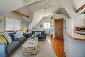Um espaço de estar tipo loft com pé-alto, ventilador de teto, cozinha com geladeira e área de estar com sofá e mesa de centro.