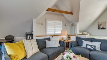 A cozy loft bedroom with a gray sofa, a yellow pillow, a small table with a lamp, and a window with blinds.