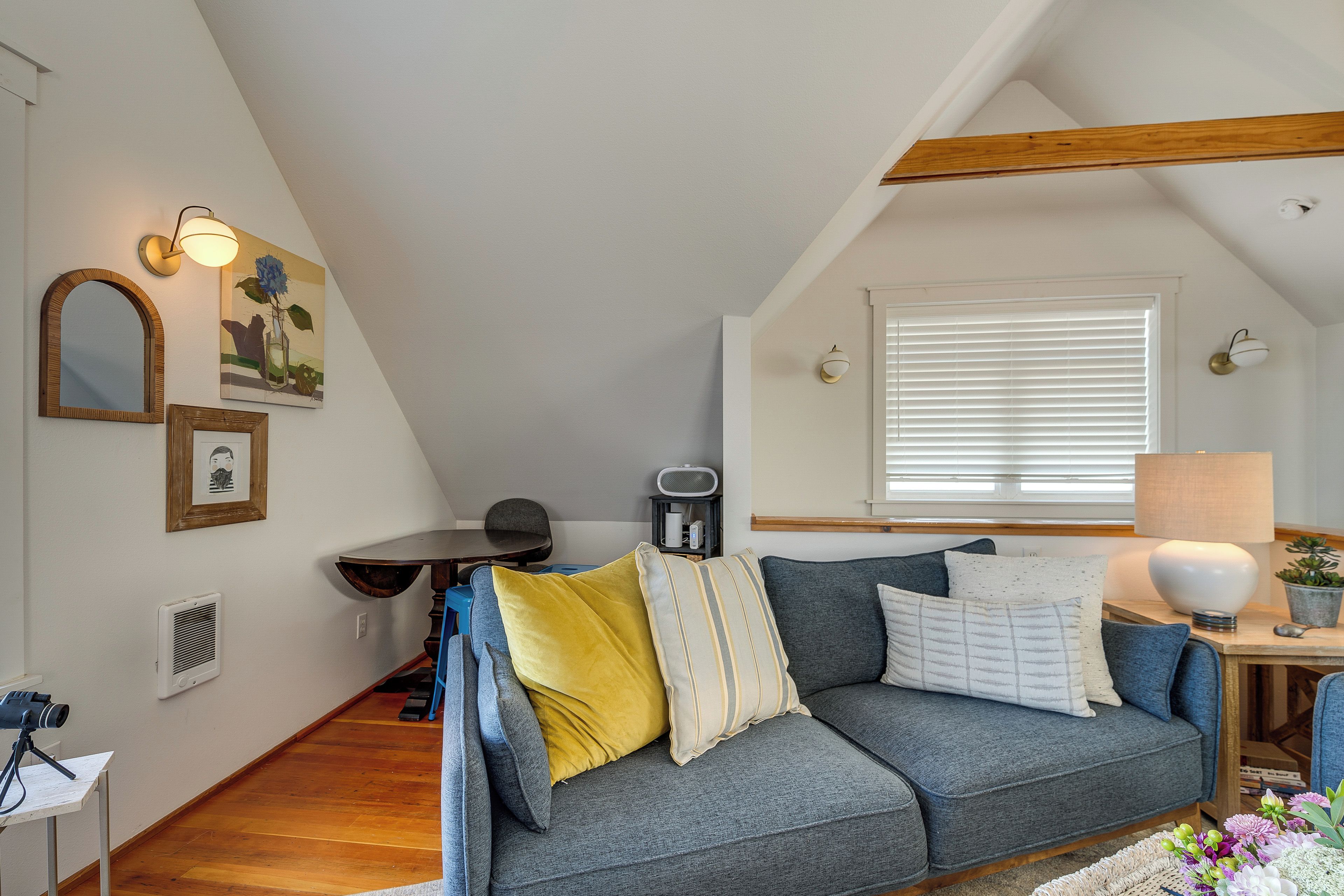 A cozy loft-style room with a grey sofa, a small table, and a window with blinds.