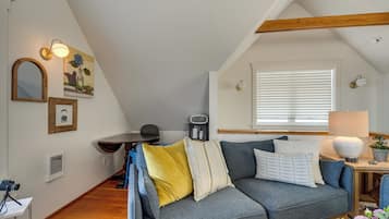 A cozy loft-style room with a grey sofa, a small table, and a window with blinds.