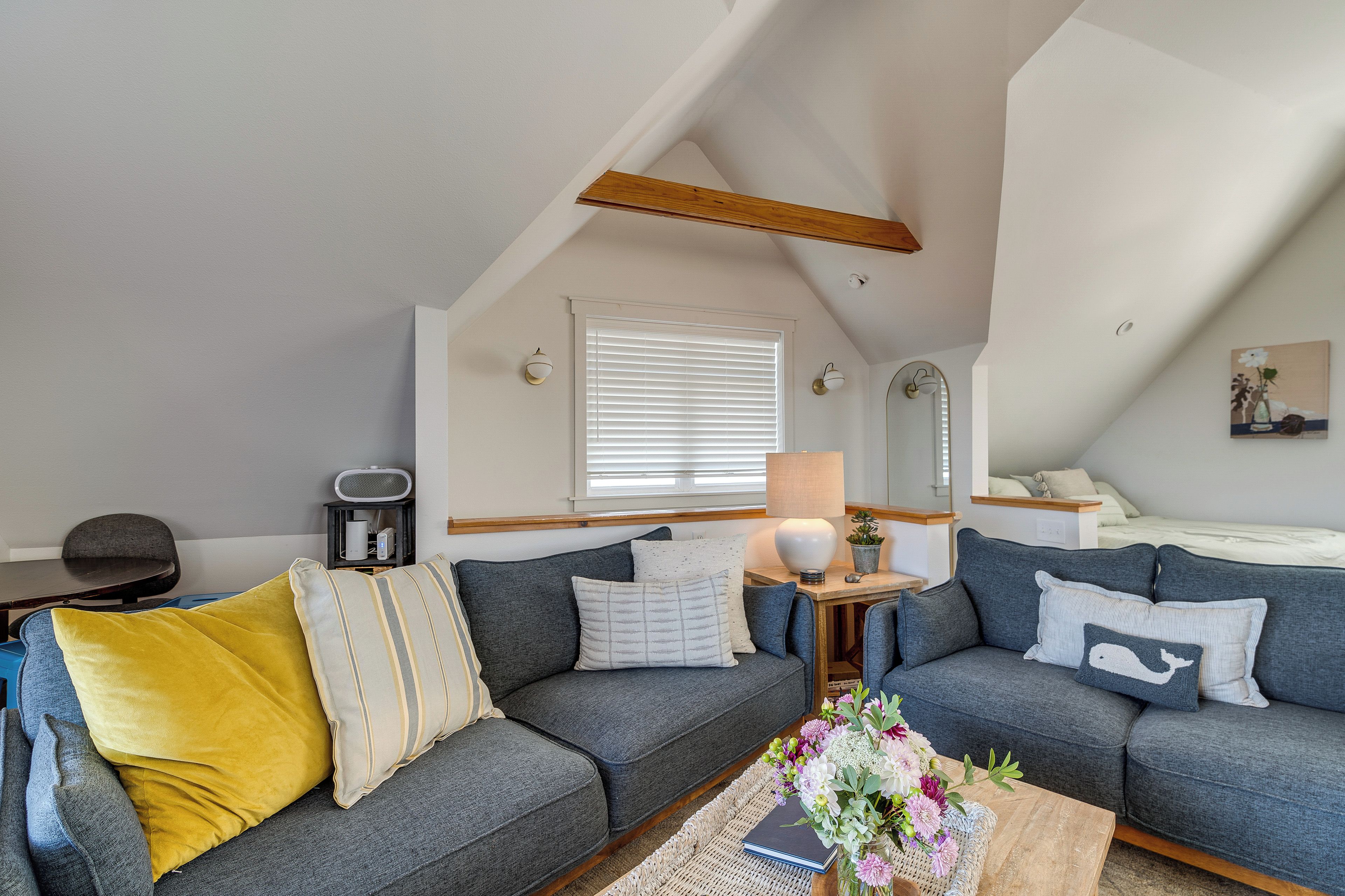 A cozy loft bedroom with a gray sofa, a yellow pillow, a small table with a lamp, and a window with blinds.