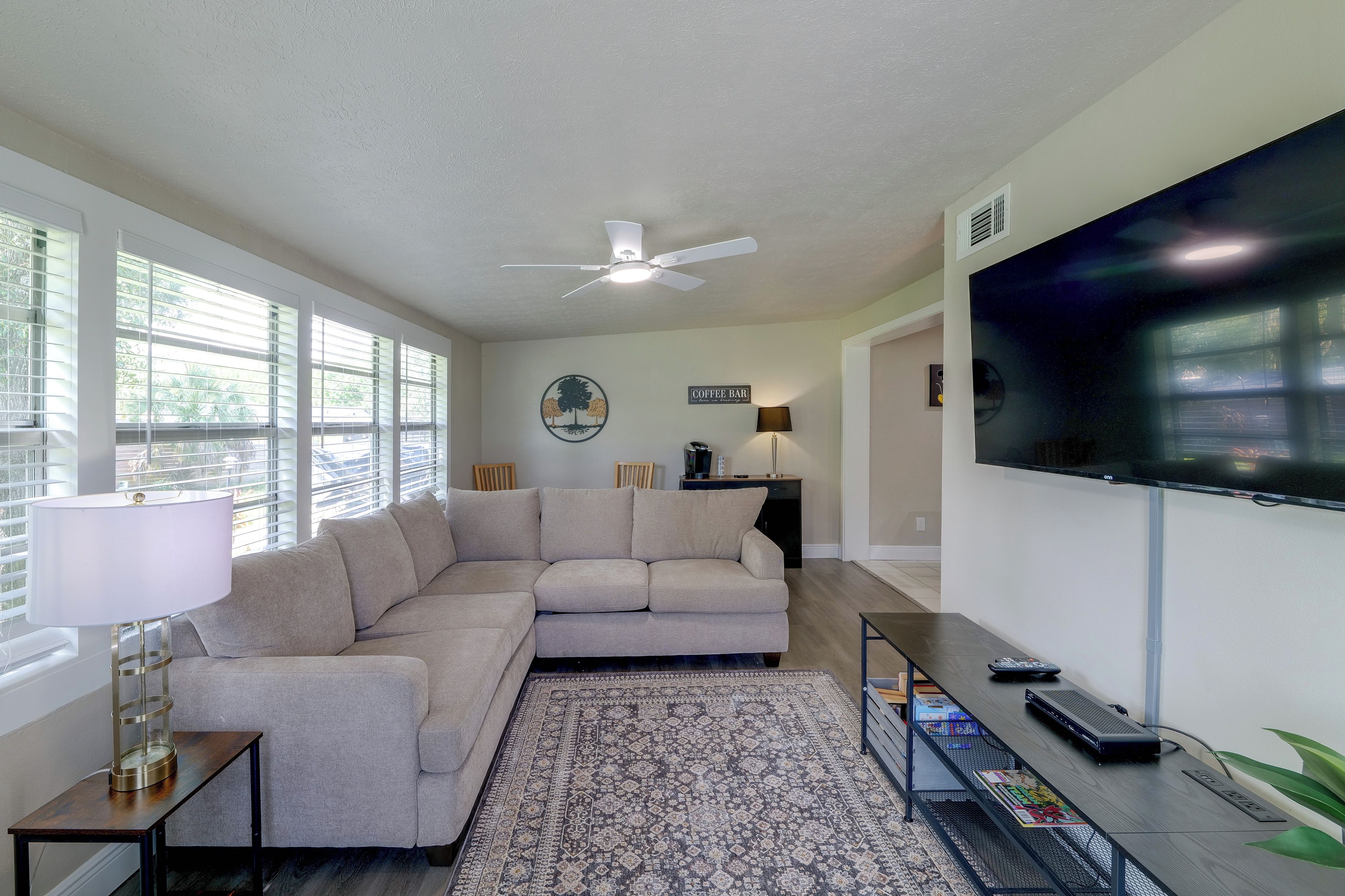 A living room with a sectional sofa, a flat-screen TV, a coffee bar sign, and a large window with blinds.