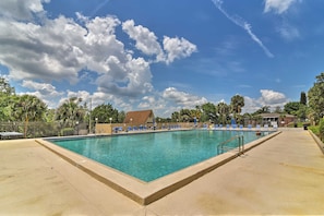 A rectangular swimming pool with clear blue water, surrounded by a concrete deck. There are lounge chairs aligned along the pool's edge, and a building with a brown roof is visible in the background. - Rum Away With Me: Resort Pool, Dock + Beach (Clermont)