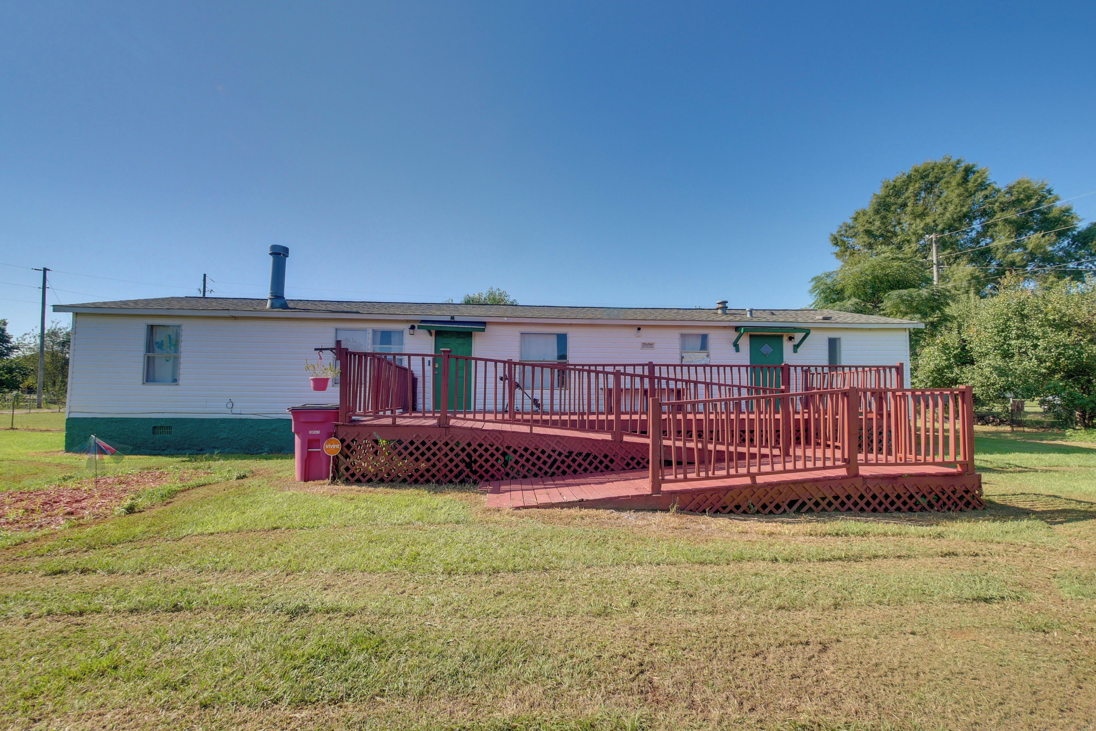 A single-story mobile home with a red deck and a green door, surrounded by a grassy yard.