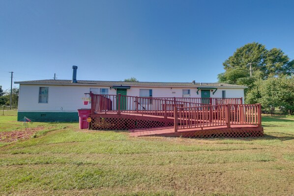 A single-story mobile home with a red deck and a green door, surrounded by a grassy yard.