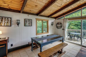 Een houten terras met een tafel en bank, een raam met uitzicht op groen, en wandlampen.