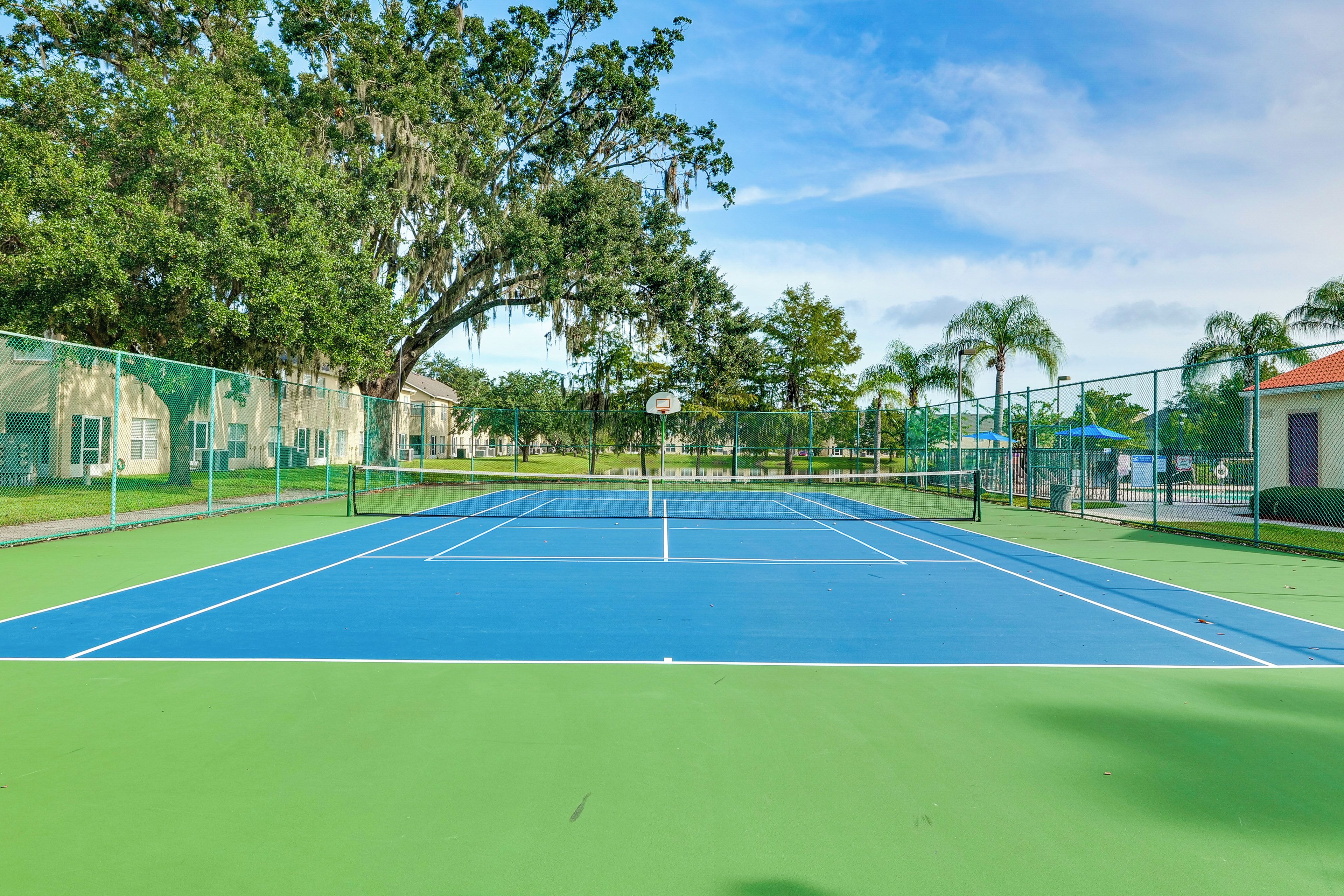 An outdoor tennis court with a net, surrounded by a green fence, trees, and a building in the background.