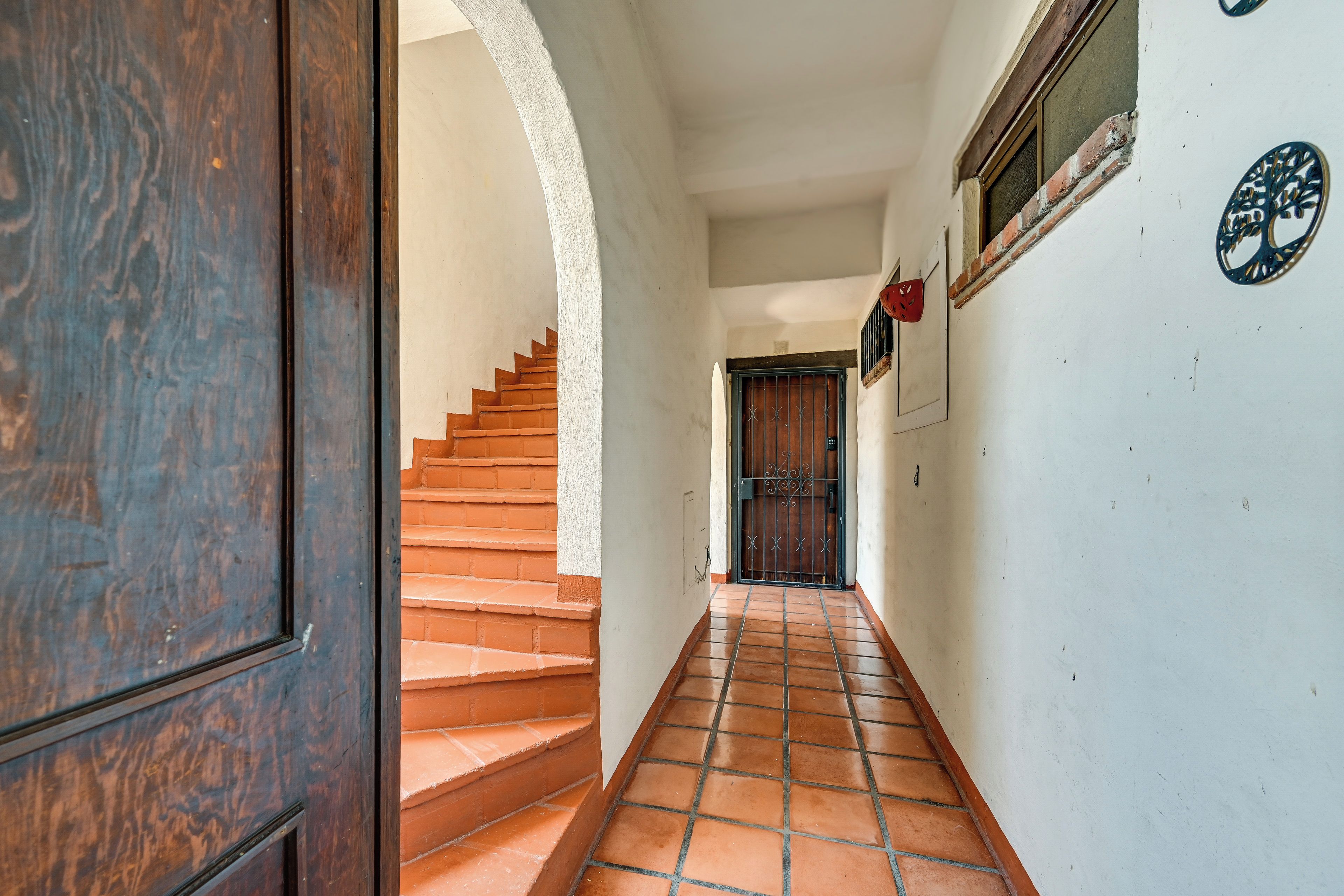 Un couloir avec une porte en bois, un escalier et un sol carrelé menant à une porte surmontée d’un portail en métal.