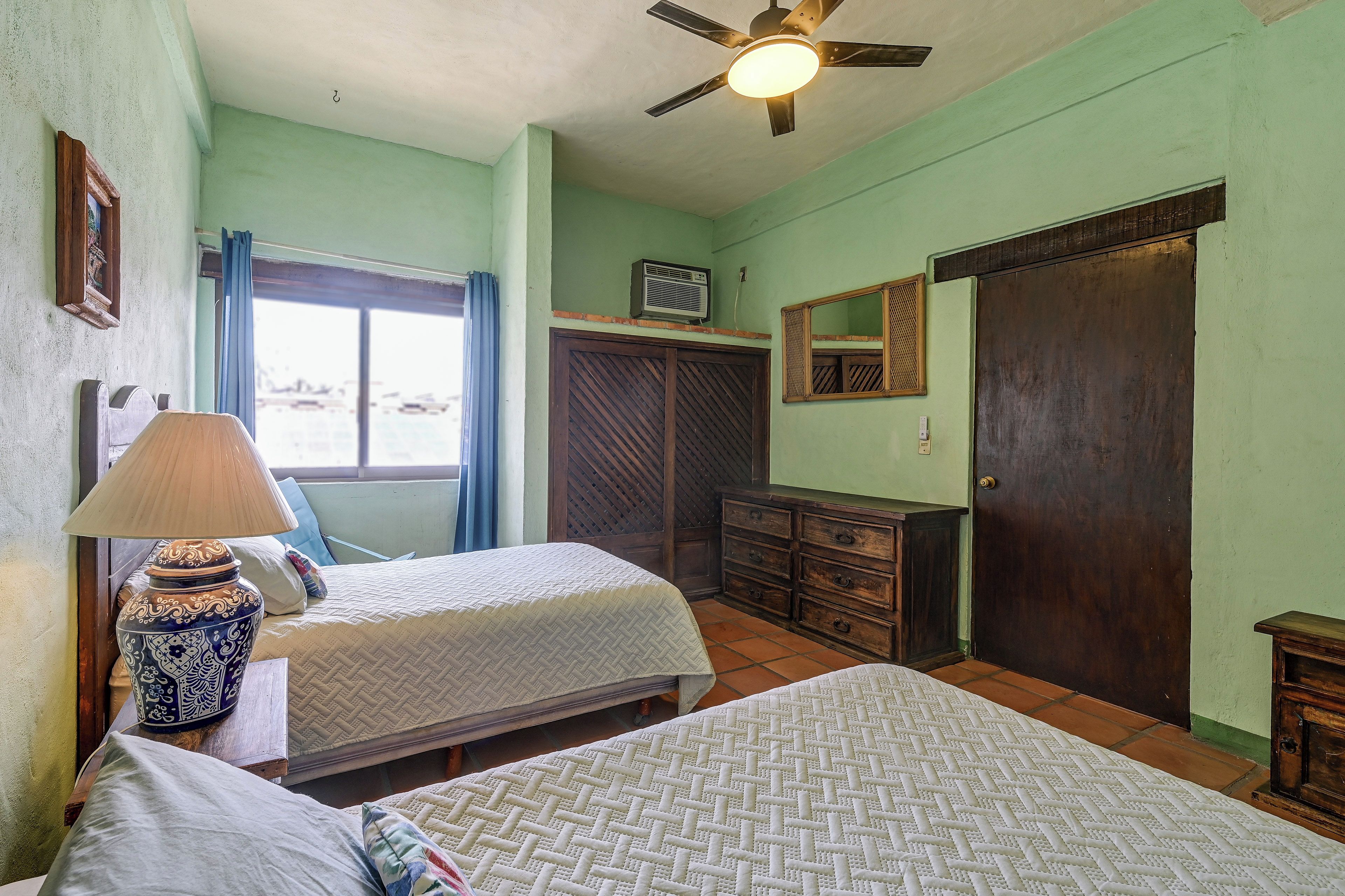 Une chambre avec deux lits, un ventilateur de plafond, une fenêtre avec des rideaux, une commode et un miroir.