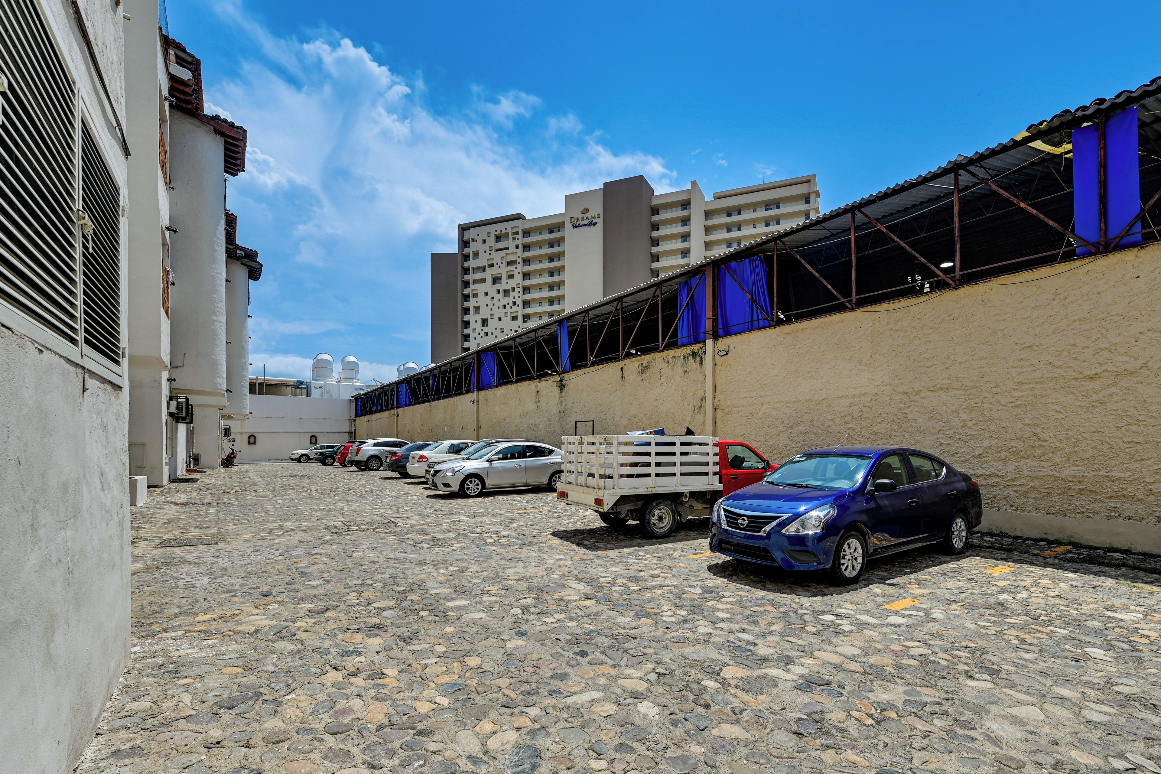 A parking area with several cars and a truck, a building with a white facade, and a blue sky.