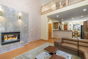 A spacious living room with a fireplace, a high ceiling, and a kitchen area visible through an open doorway. - Hot Tub & Mtn Views! Big Bear All-seasons Escape (Big Bear City)