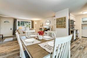 Uma área de refeições com mesa de madeira posta para quatro pessoas, cadeiras brancas e um vaso de flores vermelhas.