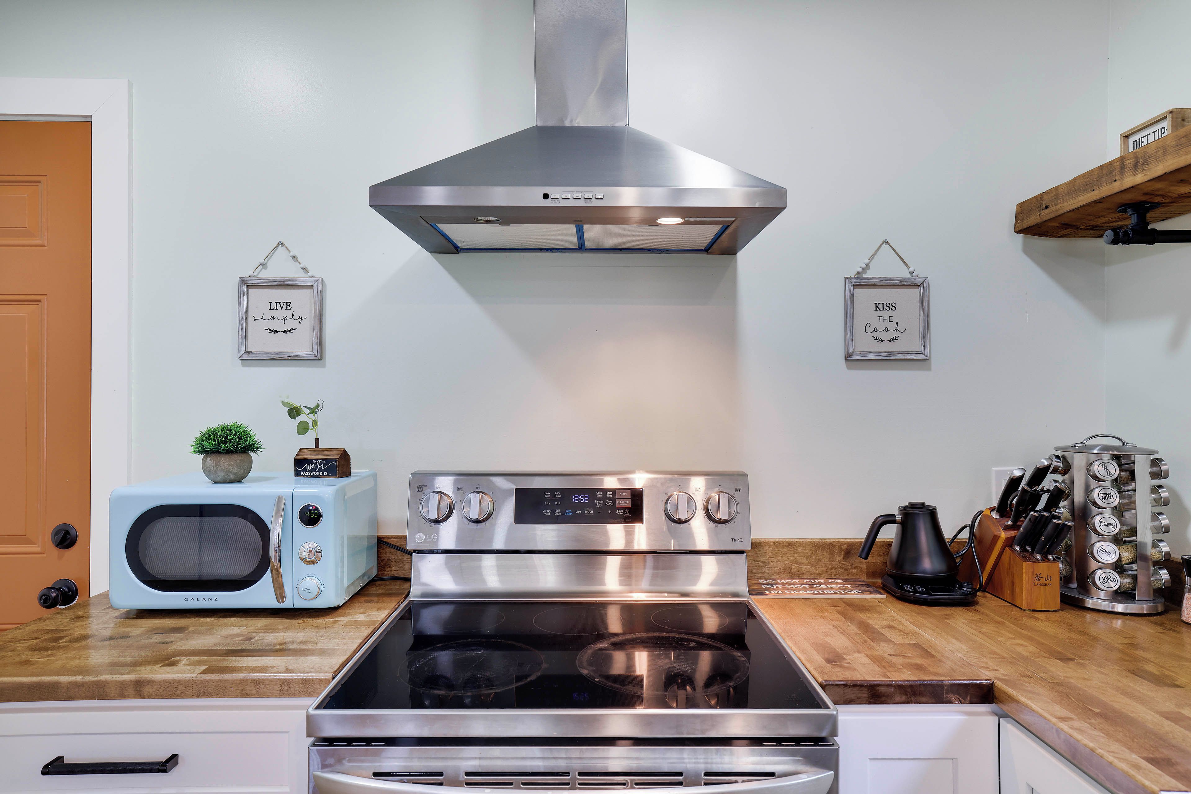 A kitchen with a stainless steel range hood, a microwave, and a stove. There are two framed signs on the wall, one reads 'LIVE simply' and the other 'KISS THE COOK'.