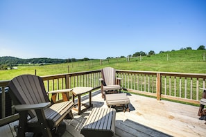 Una terraza de madera con sillas Adirondack y una mesa, con vistas a un campo cubierto de césped y una colina.