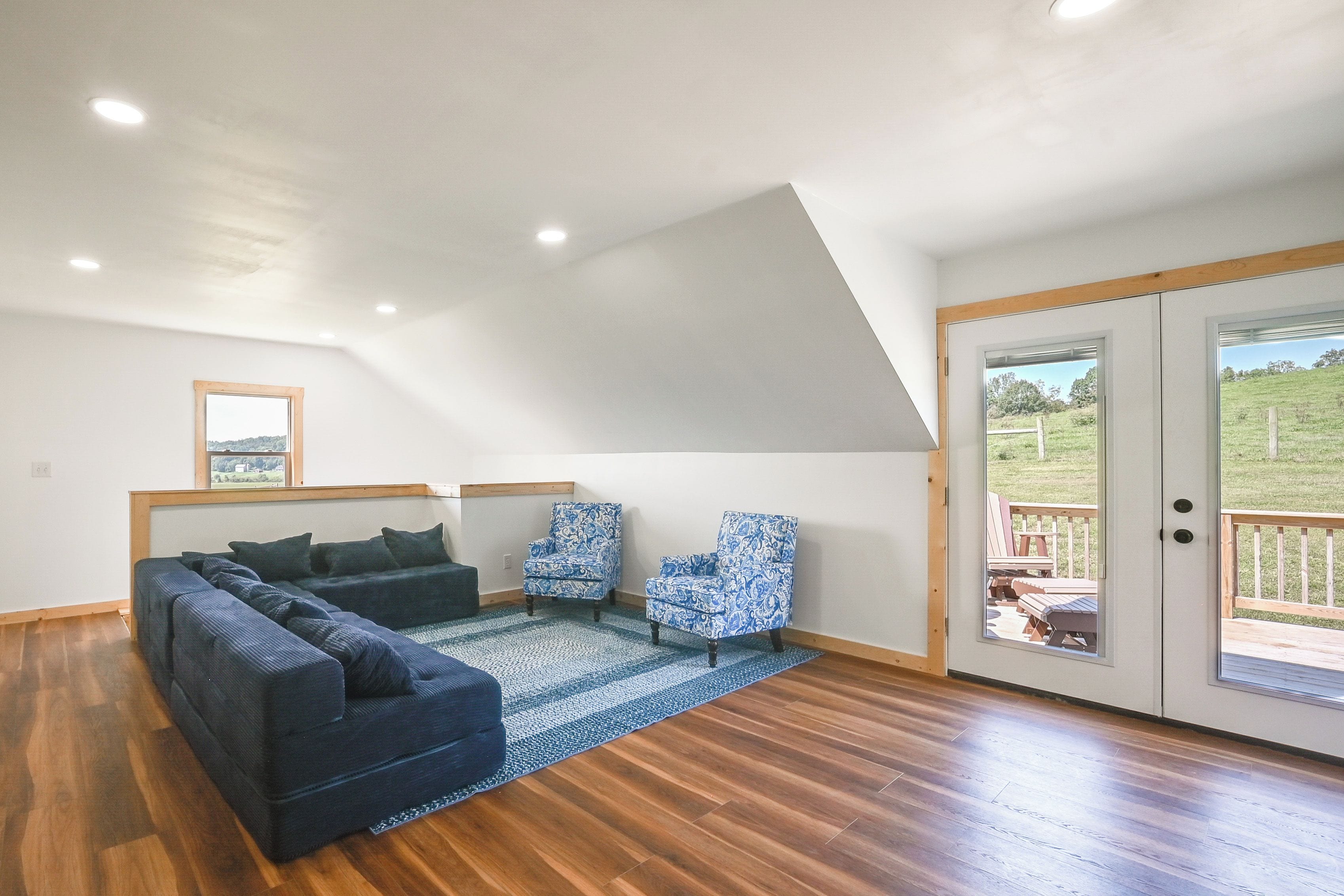 A loft-style bedroom with a sloped ceiling, a large bed, two patterned armchairs, and a view through a glass door to an outdoor deck.