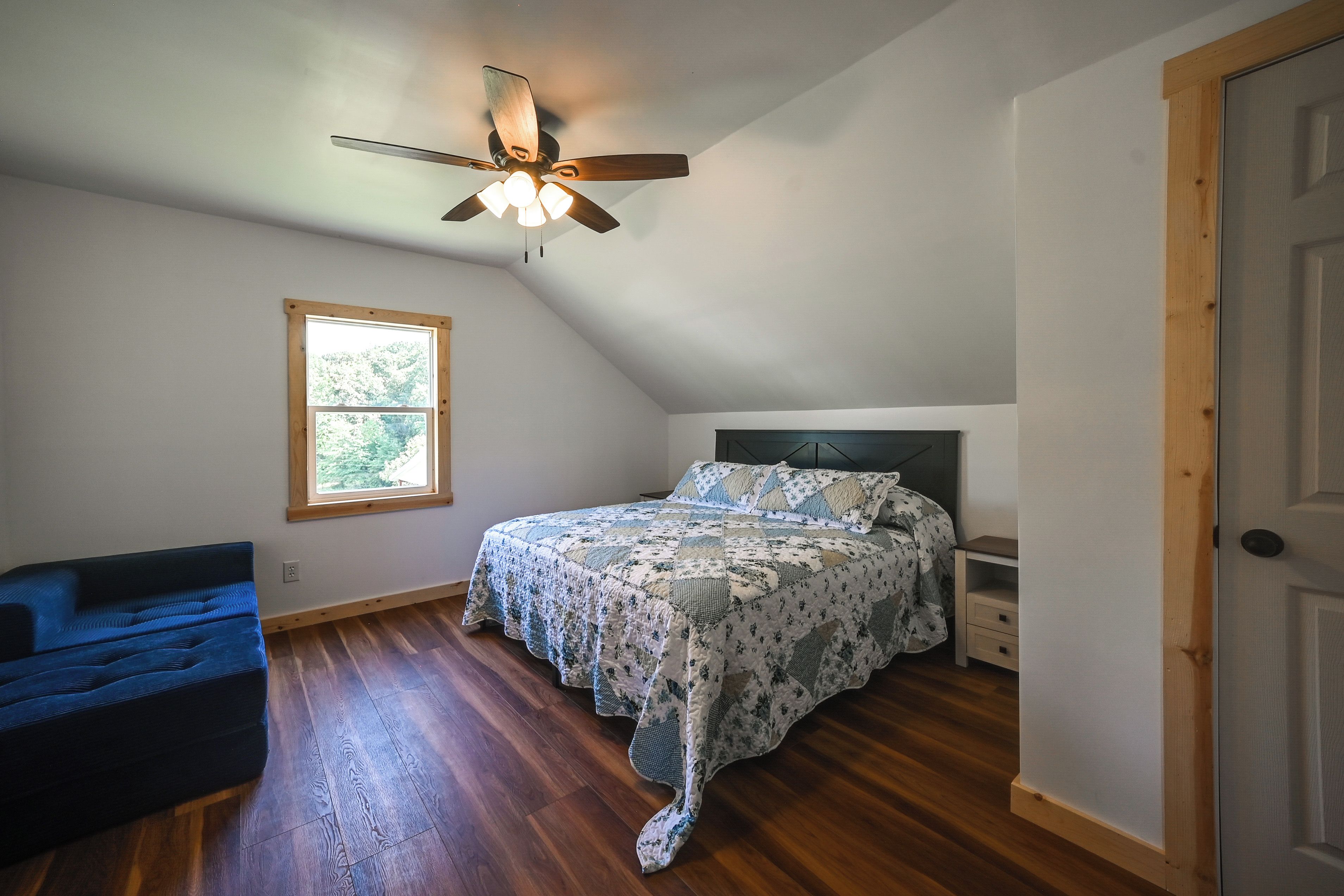 A bedroom with a sloped ceiling, a bed with a floral bedspread, a window, a blue chair, and a door.