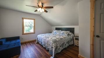 A bedroom with a sloped ceiling, a bed with a floral bedspread, a window, a blue chair, and a door.