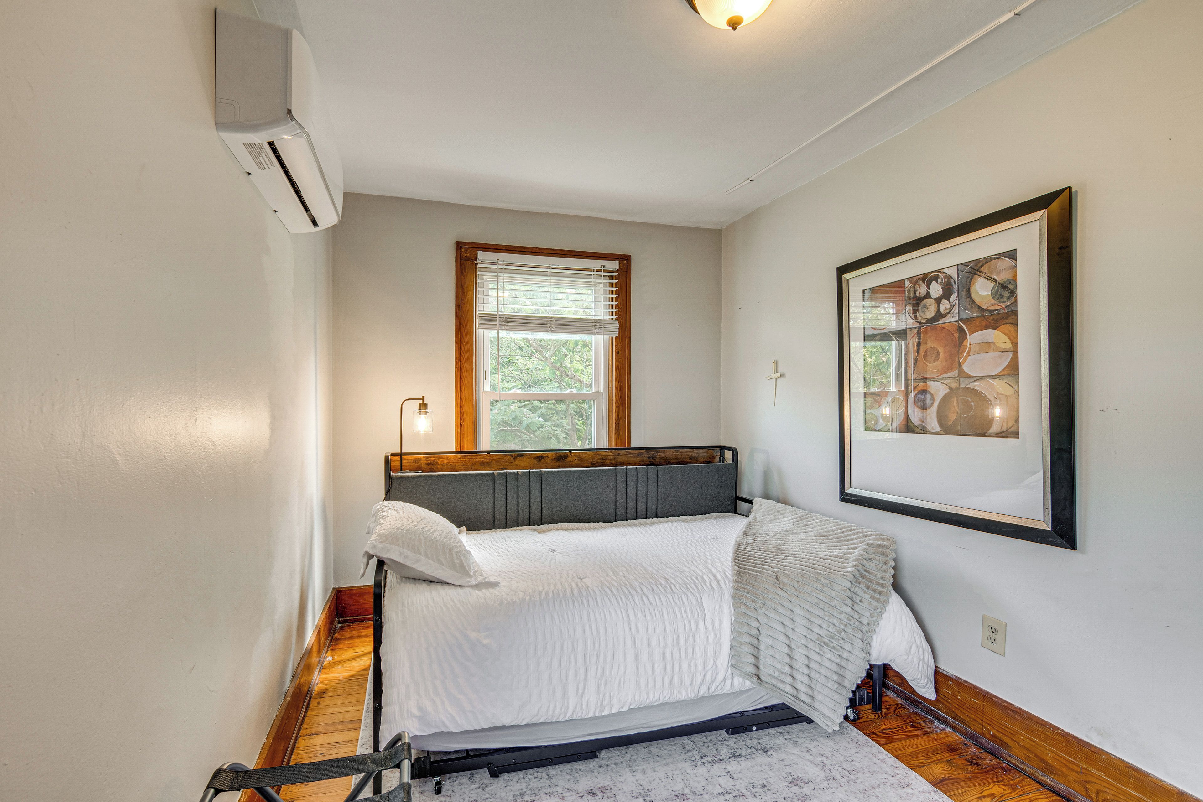 Ein Schlafzimmer mit einem Bett, einem Fenster mit Jalousien, einer Wand-Klimaanlage und einem gerahmten Bild.