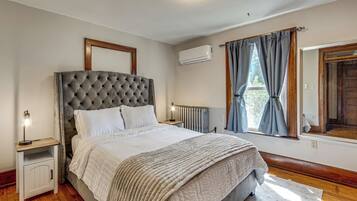 A bedroom with a tufted headboard, a bed with white bedding, a bedside table with a lamp, a window with curtains, and an air conditioning unit.