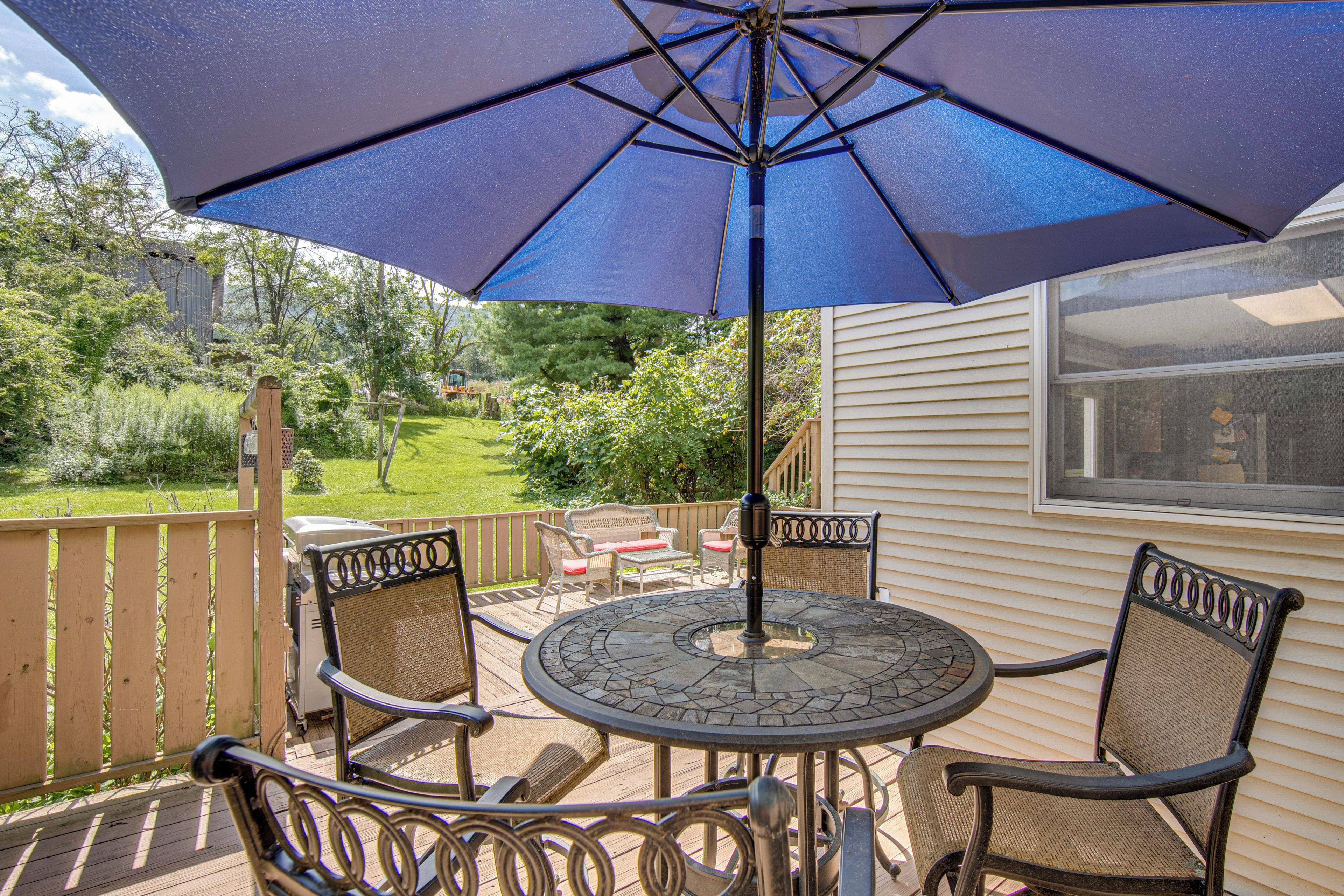 An outdoor patio with a round table and two chairs, a blue umbrella, and a view of a garden and house in the background.
