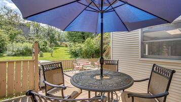 An outdoor patio with a round table and two chairs, a blue umbrella, and a view of a garden and house in the background.