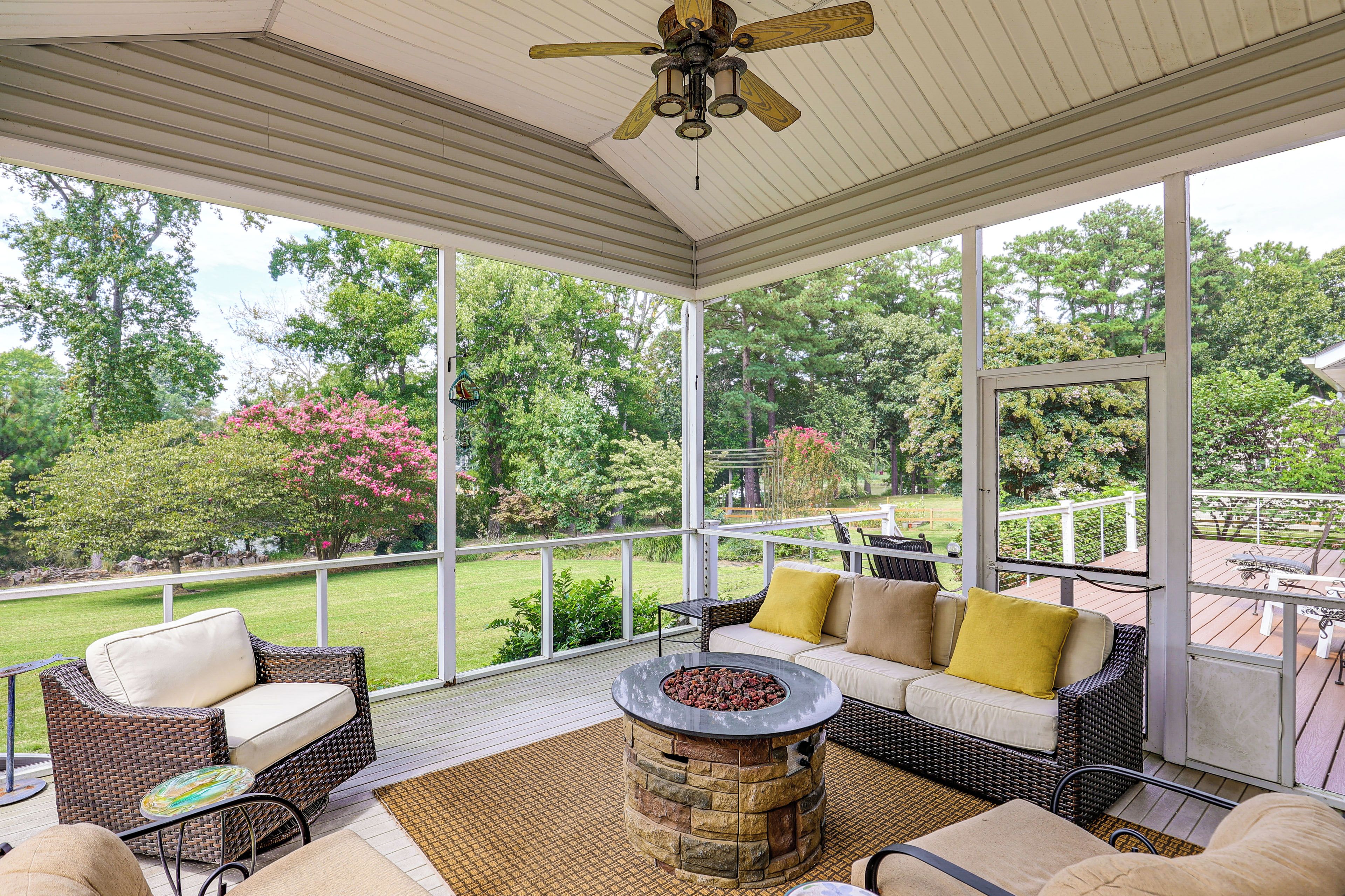 A covered outdoor seating area with wicker furniture, a stone fire pit, and a view of a garden with trees and flowers.