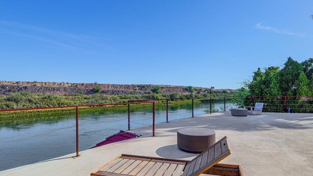 Terraza a la orilla del río con una silla de lounge de madera, un otoman redondo y una baranda de metal con vistas al río.