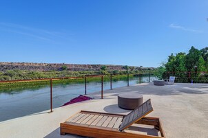 Terraza a la orilla del río con una silla de lounge de madera, un otoman redondo y una baranda de metal con vistas al río.