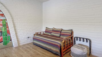 A room with a white brick wall, a wooden floor, a bed with a colorful striped bedspread, and a chair with a patterned cushion.