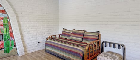 A room with a white brick wall, a wooden floor, a bed with a colorful striped bedspread, and a chair with a patterned cushion.