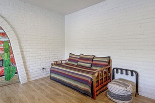 Una habitación con pared de ladrillo blanco, suelo de madera, cama con colcha a rayas de colores y silla con cojín estampado.