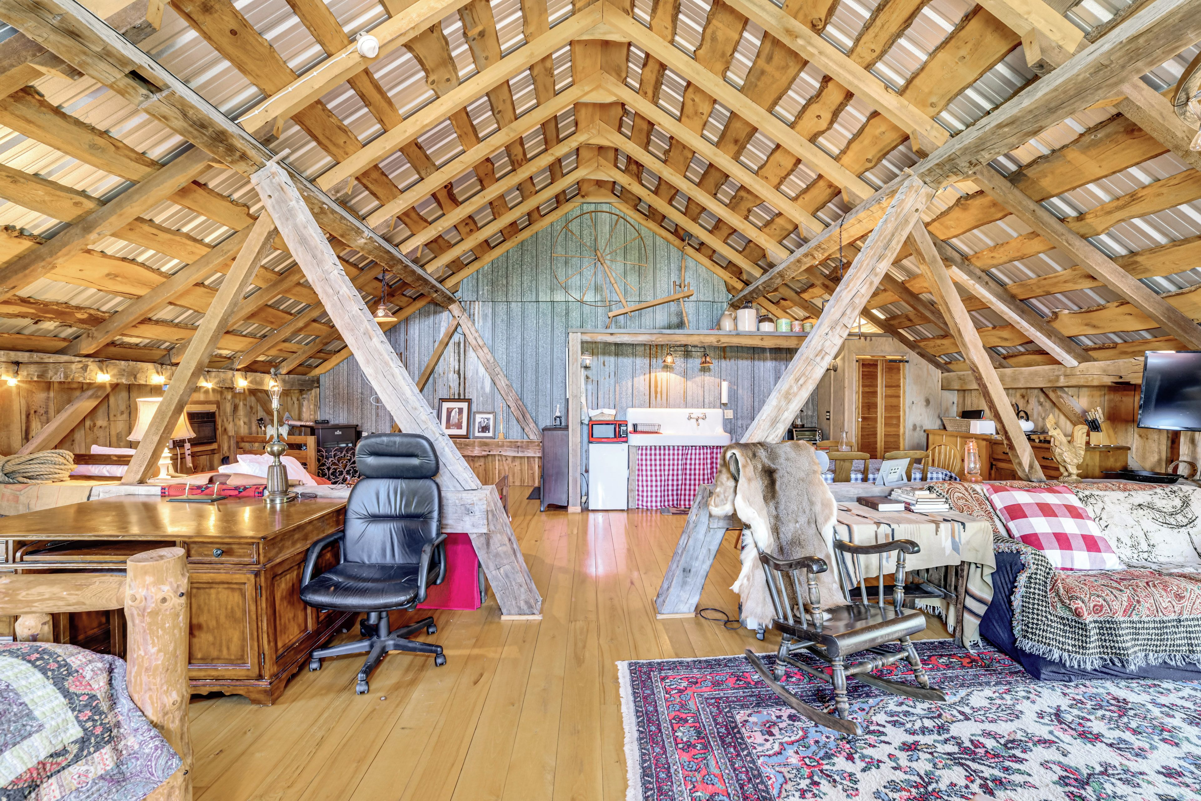 An attic room with a sloped wooden ceiling, a desk, a chair, a rocking chair, and a bed with a patterned blanket.