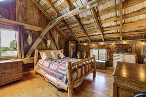 A rustic wooden room with a sloped ceiling, a bed with a patterned blanket, a dresser, and a window with curtains.