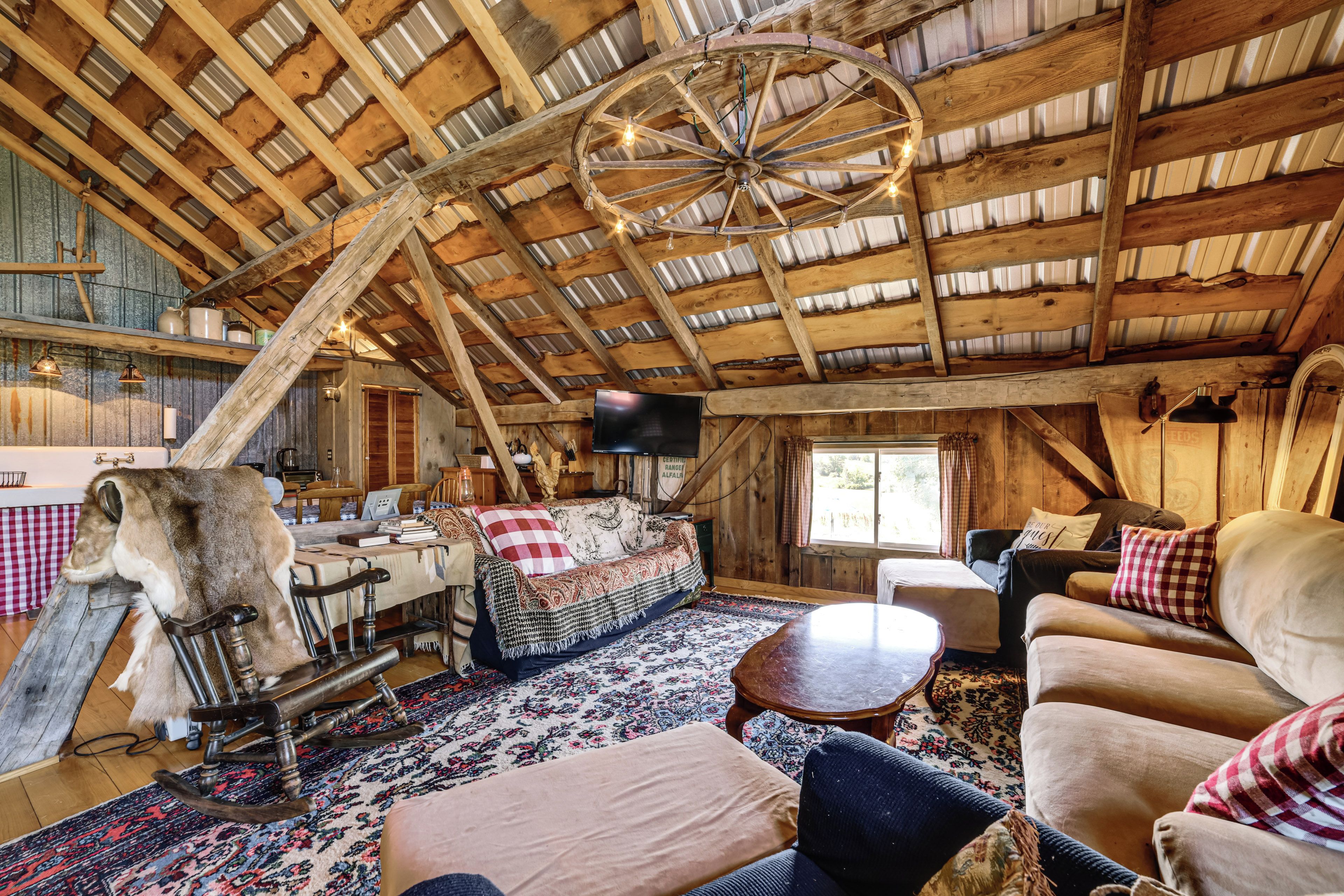 A rustic interior with a wooden ceiling, a large wheel chandelier, a sofa, a dining table, and a television.