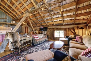 A rustic interior with a wooden ceiling, a large wheel chandelier, a sofa, a dining table, and a television.